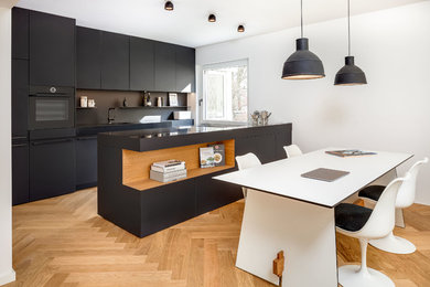 Einzeilige Moderne Wohnküche mit flächenbündigen Schrankfronten, schwarzen Schränken, Küchenrückwand in Schwarz, schwarzen Elektrogeräten, braunem Holzboden und Kücheninsel in München