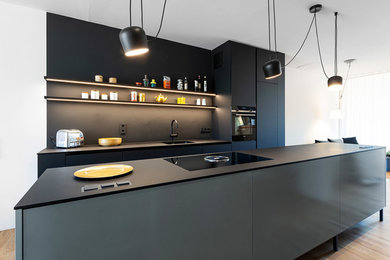 ミュンヘンにある高級な小さなモダンスタイルのおしゃれなキッチン (フラットパネル扉のキャビネット、黒いキャビネット、黒いキッチンパネル、黒い調理設備、茶色い床、黒いキッチンカウンター) の写真