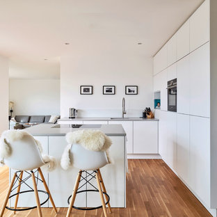 Moderne Küche in L-Form mit flächenbündigen Schrankfronten, weißen Schränken, Küchengeräten aus Edelstahl, braunem Holzboden, Kücheninsel, braunem Boden und grauer Arbeitsplatte in Berlin