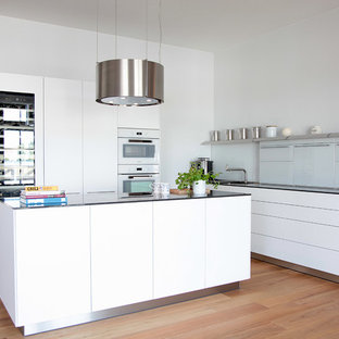 Offene, Mittelgroße Moderne Küche in L-Form mit integriertem Waschbecken, flächenbündigen Schrankfronten, weißen Schränken, Glas-Arbeitsplatte, Küchenrückwand in Weiß, Glasrückwand, weißen Elektrogeräten, braunem Holzboden, Kücheninsel, braunem Boden und schwarzer Arbeitsplatte in Hamburg