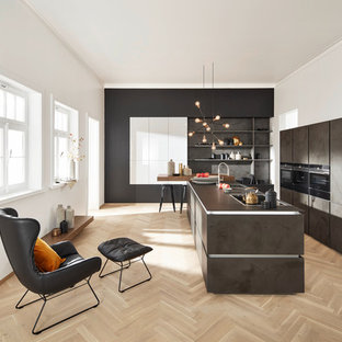 Design ideas for a contemporary galley open plan kitchen in Berlin with a drop-in sink, flat-panel cabinets, grey cabinets, grey splashback, slate splashback, light hardwood floors, with island, brown floor and black appliances.