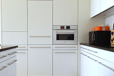 На фото: угловая кухня-гостиная среднего размера в современном стиле с плоскими фасадами, белыми фасадами, гранитной столешницей, белым фартуком, фартуком из стеклянной плитки, белой техникой, полом из керамической плитки и островом с