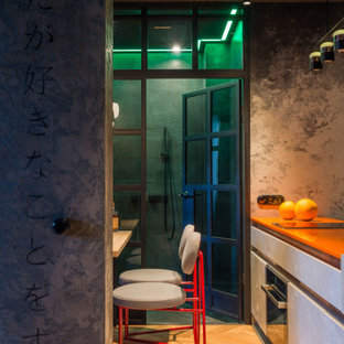 Modelo de cocina comedor de galera, urbana, pequeña, con fregadero integrado, armarios con paneles lisos, puertas de armario con efecto envejecido, encimera de cuarzo compacto, salpicadero verde, electrodomésticos negros, suelo de madera en tonos medios, península y encimeras naranjas