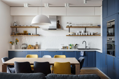 Идея дизайна: угловая кухня-гостиная в современном стиле с синими фасадами, белым фартуком, накладной мойкой, фасадами с утопленной филенкой, фартуком из плитки кабанчик, техникой из нержавеющей стали, коричневым полом, белой столешницей, паркетным полом среднего тона и двухцветным гарнитуром