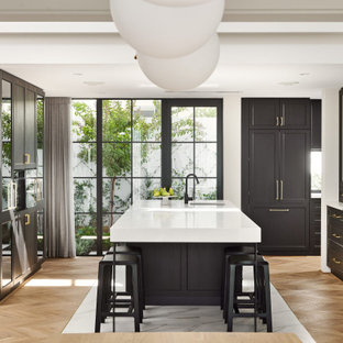 This is an example of a transitional u-shaped eat-in kitchen with black cabinets, light hardwood floors, white benchtop, an undermount sink, shaker cabinets, panelled appliances, with island and brown floor.