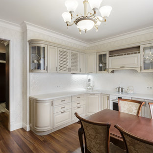 This is an example of a mid-sized traditional l-shaped eat-in kitchen in Other with an undermount sink, glass-front cabinets, beige cabinets, solid surface benchtops, white splashback, ceramic splashback, white appliances, laminate floors, no island, brown floor and white benchtop.