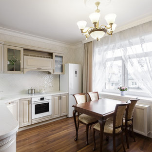 Mid-sized traditional l-shaped eat-in kitchen in Other with an undermount sink, glass-front cabinets, beige cabinets, solid surface benchtops, white splashback, ceramic splashback, white appliances, laminate floors, no island, brown floor and white benchtop.