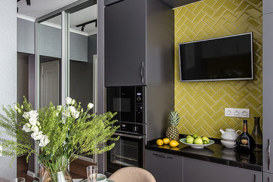 Diseño de cocina escandinava con armarios con paneles lisos, puertas de armario grises, salpicadero amarillo, salpicadero de azulejos tipo metro, electrodomésticos negros, suelo marrón y encimeras negras