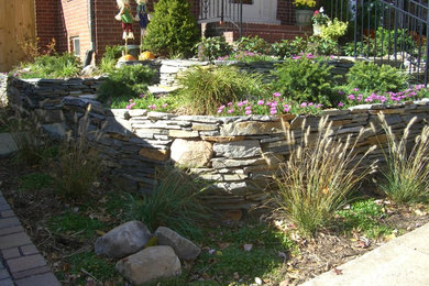 Modelo de jardín clásico renovado de tamaño medio en primavera en patio delantero con exposición total al sol, muro de contención y adoquines de piedra natural