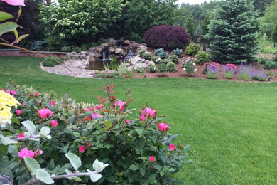 Modelo de jardín clásico de tamaño medio en verano en patio trasero con fuente, exposición reducida al sol y adoquines de piedra natural