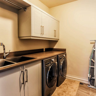Réalisation d'une buanderie linéaire champêtre dédiée et de taille moyenne avec un évier 2 bacs, un placard à porte shaker, des portes de placard grises, un plan de travail en stratifié, un mur beige, un sol en carrelage de porcelaine et des machines côte à côte.