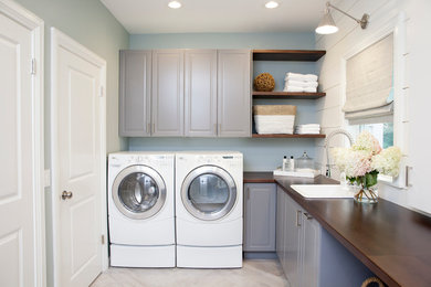 Example of a large classic l-shaped porcelain tile dedicated laundry room design in New York with raised-panel cabinets, gray cabinets, wood countertops, blue walls, a side-by-side washer/dryer, a drop-in sink and brown countertops