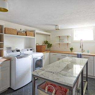 Idéer för stora vintage l-formade brunt tvättstugor enbart för tvätt, med en nedsänkt diskho, skåp i shakerstil, vita skåp, träbänkskiva, vita väggar, skiffergolv, en tvättmaskin och torktumlare bredvid varandra och grått golv