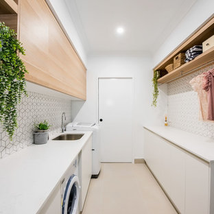 Immagine di una sala lavanderia minimal di medie dimensioni con lavello sottopiano, ante lisce, pareti bianche, lavatrice e asciugatrice affiancate, top bianco, ante in legno chiaro, top in granito, paraspruzzi bianco, paraspruzzi con piastrelle in ceramica, pavimento con piastrelle in ceramica e pavimento bianco