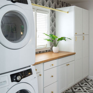 Immagine di una sala lavanderia tradizionale di medie dimensioni con ante in stile shaker, ante bianche, top in legno, pareti bianche, pavimento con piastrelle in ceramica, lavatrice e asciugatrice a colonna, pavimento nero, top marrone e carta da parati