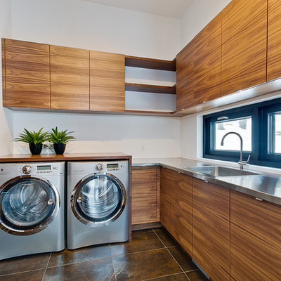 Example of a trendy l-shaped brown floor dedicated laundry room design in Montreal with a drop-in sink, flat-panel cabinets, medium tone wood cabinets, stainless steel countertops, white walls, a side-by-side washer/dryer and gray countertops