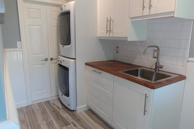 Example of a mid-sized beach style galley porcelain tile and gray floor dedicated laundry room design in Philadelphia with a single-bowl sink, shaker cabinets, white cabinets, wood countertops, gray walls, a stacked washer/dryer and brown countertops