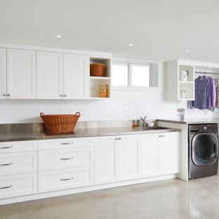 Cette photo montre une très grand buanderie linéaire chic multi-usage avec un évier intégré, un placard avec porte à panneau encastré, des portes de placard blanches, un plan de travail en inox, un mur blanc, béton au sol, des machines côte à côte et un sol gris.