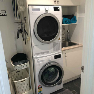 Foto på en liten beige linjär tvättstuga enbart för tvätt, med en enkel diskho, vita skåp, laminatbänkskiva, vitt stänkskydd, stänkskydd i porslinskakel, vita väggar, klinkergolv i porslin, en tvättpelare och grått golv