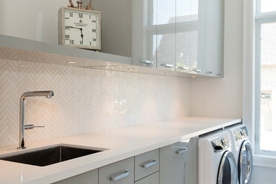 Immagine di una sala lavanderia contemporanea con lavello sottopiano, ante lisce, ante grigie, pareti bianche, lavatrice e asciugatrice affiancate, pavimento bianco e top bianco