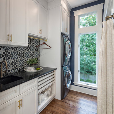 Inspiration for a transitional single-wall dark wood floor and brown floor dedicated laundry room remodel in Cincinnati with an undermount sink, recessed-panel cabinets, white cabinets, blue walls, a stacked washer/dryer and black countertops