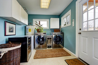 Modelo de cuarto de lavado en L clásico de tamaño medio con armarios con paneles lisos, puertas de armario blancas, paredes azules, suelo laminado, lavadora y secadora juntas y suelo beige