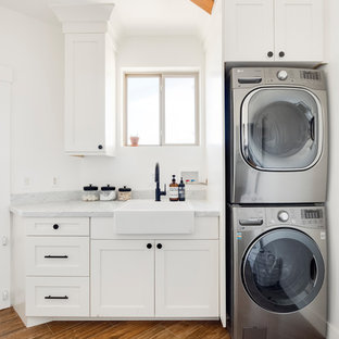 Exemple d'une petit buanderie linéaire nature dédiée avec un évier de ferme, un placard à porte shaker, des portes de placard blanches, un plan de travail en marbre, un mur blanc, un sol en carrelage de porcelaine, des machines superposées, un sol marron et un plan de travail gris.