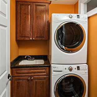 Inspiration för små amerikanska linjära svart tvättstugor enbart för tvätt, med en undermonterad diskho, luckor med upphöjd panel, skåp i mörkt trä, granitbänkskiva, orange väggar, mellanmörkt trägolv, en tvättpelare och brunt golv
