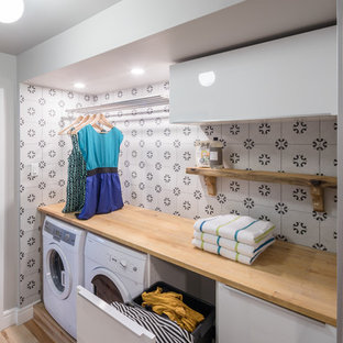 Idéer för att renovera en liten funkis beige linjär beige tvättstuga enbart för tvätt, med släta luckor, vita skåp, träbänkskiva, grå väggar, ljust trägolv och en tvättmaskin och torktumlare bredvid varandra