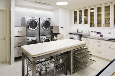 ニューヨークにある巨大なトランジショナルスタイルのおしゃれな洗濯室 (コの字型、アンダーカウンターシンク、ガラス扉のキャビネット、白いキャビネット、ステンレスカウンター、白い壁、上下配置の洗濯機・乾燥機) の写真