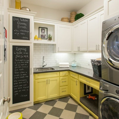 Elegant l-shaped laundry room photo in Dallas with an undermount sink, shaker cabinets, yellow cabinets, white walls, a stacked washer/dryer and gray countertops