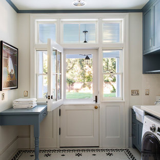 Exemple d'une grand buanderie linéaire chic avec un évier de ferme, un placard à porte shaker, des portes de placard bleues, un mur blanc, des machines côte à côte, un plan de travail en quartz, un sol en carrelage de céramique, un sol multicolore et un plan de travail blanc.