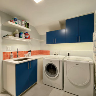 Inspiration för mellanstora moderna vitt grovkök, med en allbänk, släta luckor, blå skåp, laminatbänkskiva, orange stänkskydd, stänkskydd i keramik, vita väggar, klinkergolv i porslin, en tvättmaskin och torktumlare bredvid varandra och grått golv