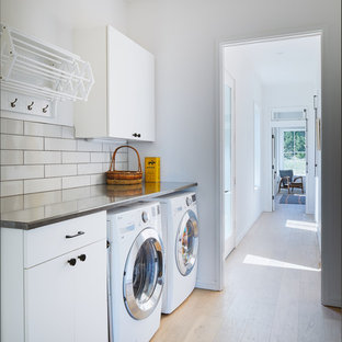 Idéer för att renovera en minimalistisk grå linjär grått tvättstuga enbart för tvätt, med släta luckor, vita skåp, bänkskiva i rostfritt stål, vita väggar, ljust trägolv, en tvättmaskin och torktumlare bredvid varandra och beiget golv
