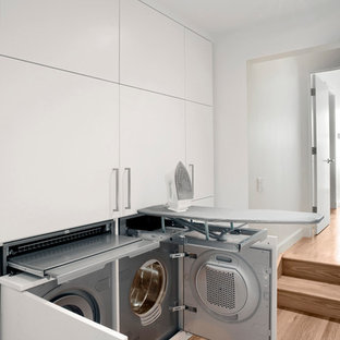 Inredning av en modern liten liten tvättstuga, med släta luckor, vita skåp, vita väggar, mellanmörkt trägolv och en tvättmaskin och torktumlare bredvid varandra