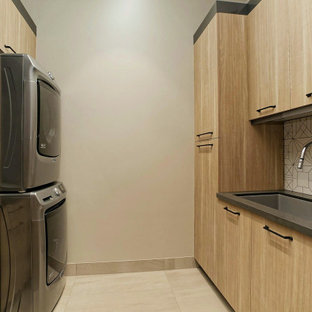 Idée de décoration pour une buanderie parallèle chalet dédiée avec un évier encastré, un placard à porte plane, des portes de placard en bois clair, une crédence multicolore, un mur beige, des machines superposées, un sol beige et un plan de travail gris.