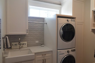Example of a mid-sized transitional porcelain tile and brown floor utility room design in Chicago with a farmhouse sink, recessed-panel cabinets, white cabinets, quartz countertops, gray walls and a stacked washer/dryer