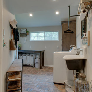 Aménagement d'une petit buanderie campagne multi-usage avec un évier de ferme, un placard à porte shaker, des portes de placard marrons, un plan de travail en granite, un mur gris, un sol en brique, des machines côte à côte, un sol multicolore et un plan de travail noir.