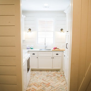 Ispirazione per una sala lavanderia country di medie dimensioni con lavello sottopiano, ante con riquadro incassato, ante bianche, pareti bianche, pavimento in mattoni, lavatrice e asciugatrice affiancate, pavimento beige, top in cemento e top grigio