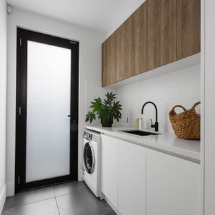 Cette photo montre une buanderie linéaire tendance avec un évier encastré, un placard à porte plane, des portes de placard blanches, une crédence blanche, un mur blanc, le lave-linge et le sèche-linge forment un seul appareil électroménager, un sol gris et un plan de travail blanc.