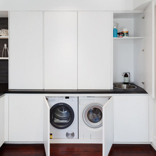 Exempel på en modern svarta svart tvättstuga, med en enkel diskho, vita skåp, granitbänkskiva, vita väggar, mörkt trägolv och tvättmaskin och torktumlare byggt in i ett skåp