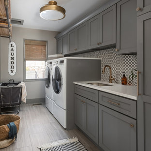 Modelo de cuarto de lavado lineal, clásico renovado, de tamaño medio, con fregadero bajoencimera, armarios estilo shaker, puertas de armario grises, encimera de cuarzo compacto, salpicadero multicolor, salpicadero de azulejos de cemento, paredes grises, suelo de baldosas de porcelana, lavadora y secadora juntas, suelo gris y encimeras blancas