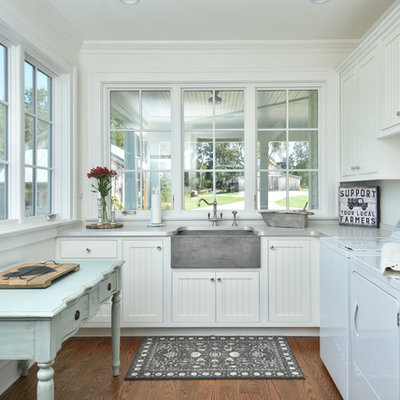 Example of a cottage dark wood floor and brown floor utility room design in Other with a farmhouse sink, recessed-panel cabinets, white cabinets, white walls, a side-by-side washer/dryer, laminate countertops and gray countertops