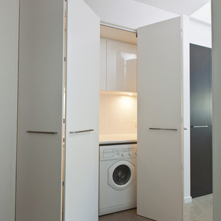 Modern inredning av en liten linjär tvättstuga enbart för tvätt, med släta luckor, vita skåp, laminatbänkskiva, vita väggar, betonggolv och en tvättmaskin och torktumlare bredvid varandra
