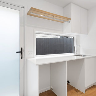 Inspiration för linjära vitt tvättstugor enbart för tvätt, med en nedsänkt diskho, släta luckor, vita skåp, fönster som stänkskydd, en tvättmaskin och torktumlare bredvid varandra och brunt golv