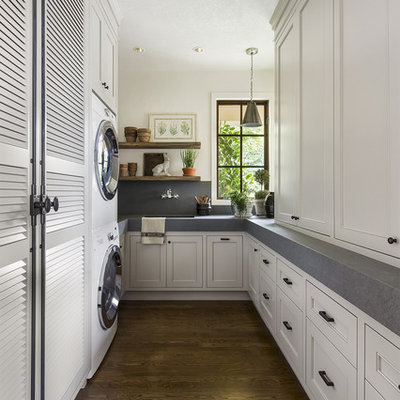Example of a classic u-shaped dark wood floor laundry room design in Portland with an undermount sink, recessed-panel cabinets, white cabinets, white walls, a stacked washer/dryer and gray countertops