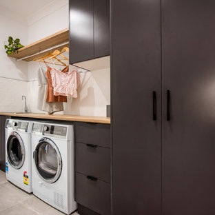 This is an example of a mid-sized contemporary single-wall dedicated laundry room in Other with a drop-in sink, flat-panel cabinets, grey cabinets, wood benchtops, white splashback, ceramic splashback, white walls, ceramic floors, a side-by-side washer and dryer and grey floor.