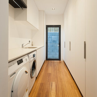 Inspiration pour une grand buanderie parallèle design dédiée avec un évier encastré, un placard à porte plane, des portes de placard blanches, un plan de travail en stratifié, un mur blanc, un sol en bois clair et des machines côte à côte.