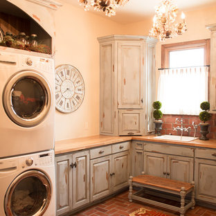 Lantlig inredning av en mellanstor beige l-formad beige tvättstuga enbart för tvätt, med en nedsänkt diskho, luckor med upphöjd panel, skåp i slitet trä, träbänkskiva, beige väggar, tegelgolv, en tvättpelare och rött golv
