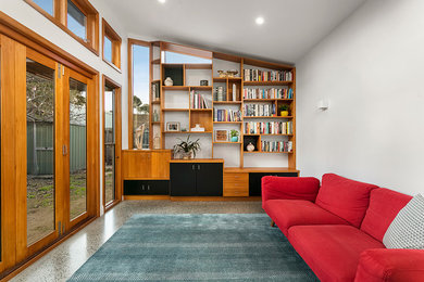 Modelo de biblioteca en casa cerrada moderna grande sin chimenea con paredes blancas, suelo de cemento, televisor retractable y suelo gris